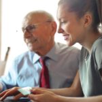 Smart Assistants - Young woman in casual clothes helping senior man in formal shirt with paying credit card in Internet using laptop while sitting at table