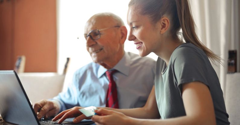 Smart Assistants - Young woman in casual clothes helping senior man in formal shirt with paying credit card in Internet using laptop while sitting at table