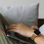Smart Home Hub - Side view of crop unrecognizable female freelancer in casual clothes and smartwatch lying on comfortable sofa and working remotely on laptop