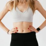 Wearables - Faceless slim anonymous blond female in sports bra and black leggings in wearable bracelet showing perfect belly on white background while standing with hands on waist