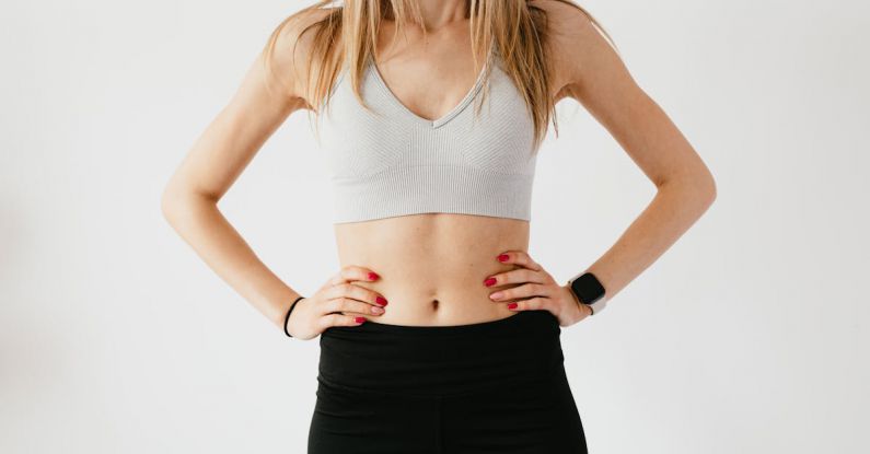 Wearables - Faceless slim anonymous blond female in sports bra and black leggings in wearable bracelet showing perfect belly on white background while standing with hands on waist